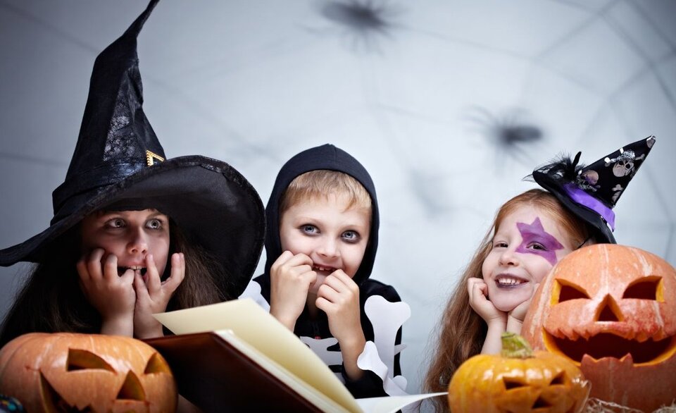 Three children in Halloween costumes sit at a table with carved pumpkins. One wears a black witch hat and robe, another is dressed as a skeleton, and the third has a purple star painted around one eye and a small witch hat.